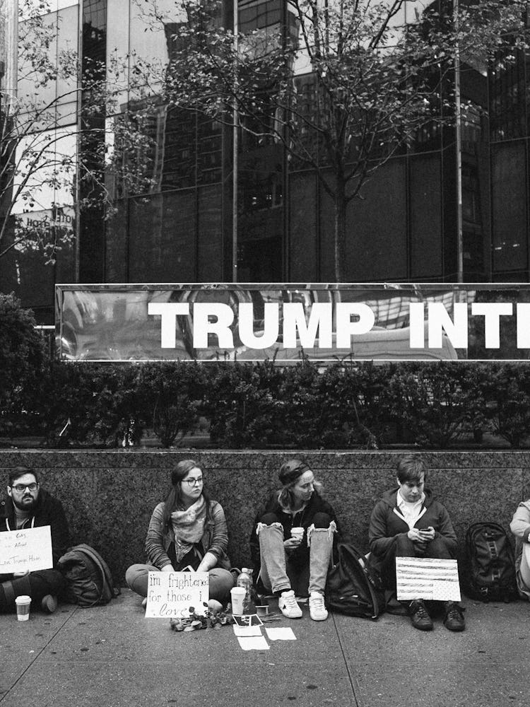Five activists protesting and sitting in front of a short wall with protest signs next to a Trump bu...