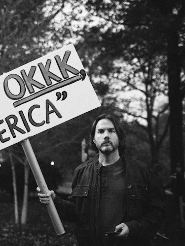 A man in a black jacket holding a protest sign with the text 'Not OKKK, America'