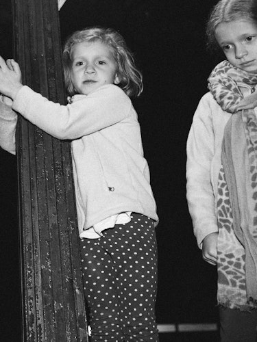 A little girl in a white sweater and polka dot pants clinging onto a wooden installation