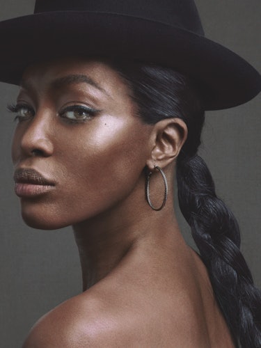 A portrait of Naomi Campbell with braided hair, a black hat and hoop earrings