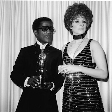 Sammy Davis in a jacket and shirt and Barbra Streisand in a gown with sequins and long necklace