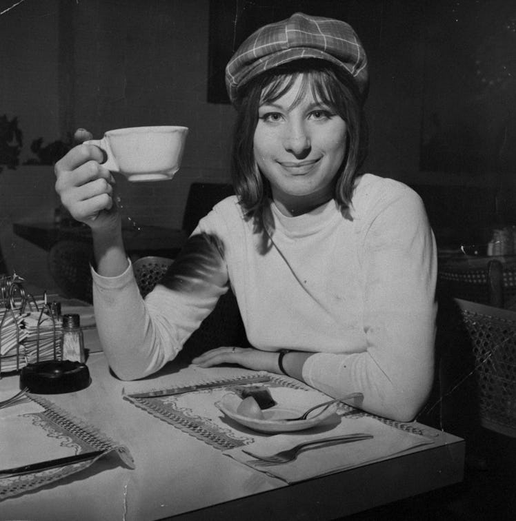 Barbra Streisand in a turtleneck and a checked hat sitting, smiling and holding a white cup in 1963