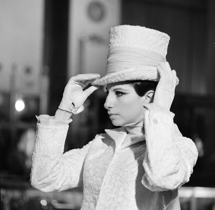 Barbra Streisand putting on a white hat while wearing matching white gloves and a white coat 