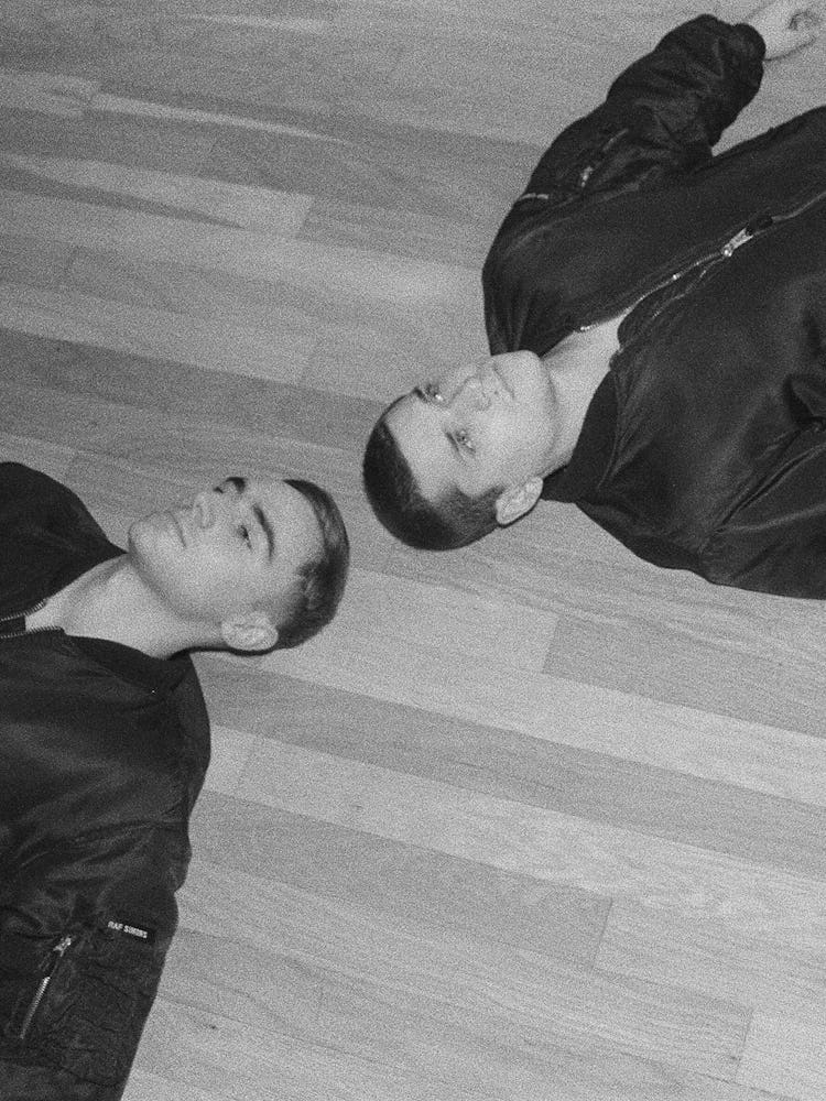 A black and white photo of two men lying on the floor in the same black bomber jacket,  one has on b...