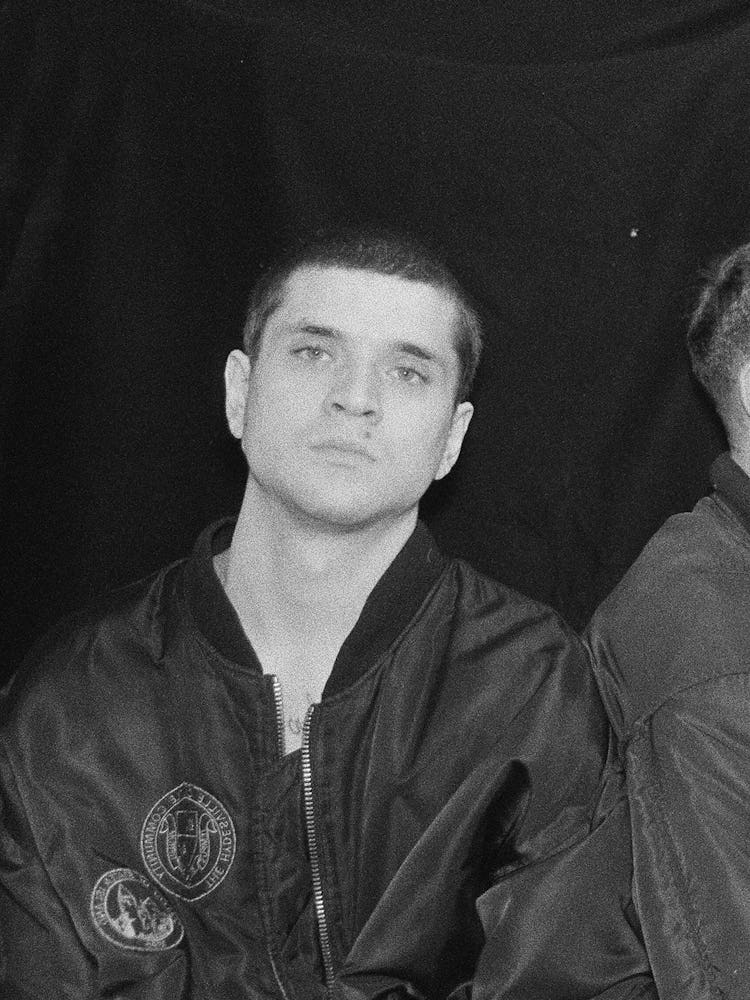 A black and white photo of men in matching black bomber jackets, one is looking straight while the o...
