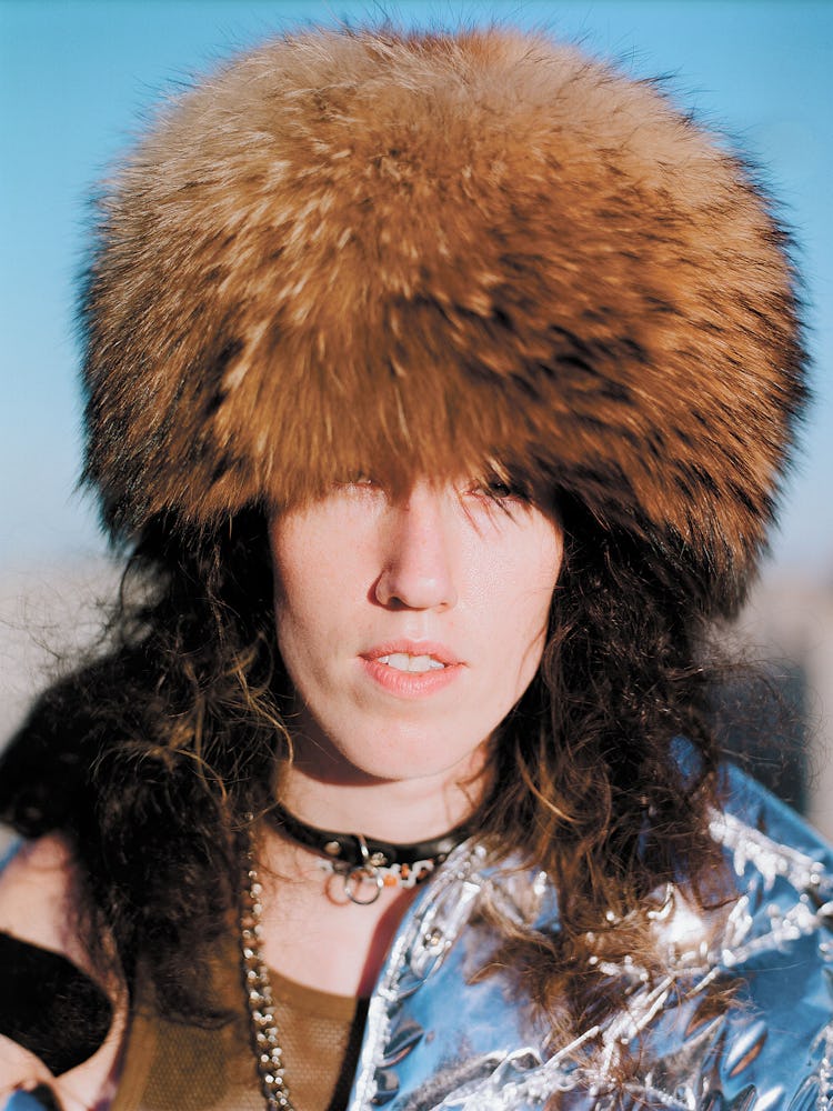 A woman wearing a light brown fur hat, grey metallic jacket and black leather choker 