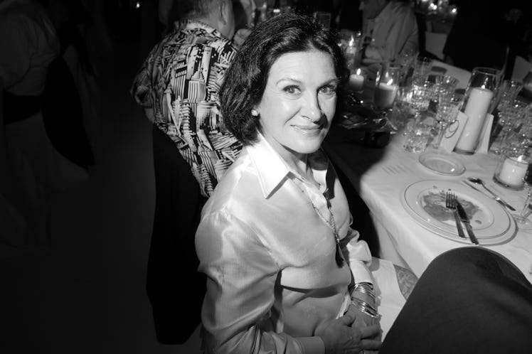 Paloma Picasso sitting at a table and smiling while wearing a white satin shirt