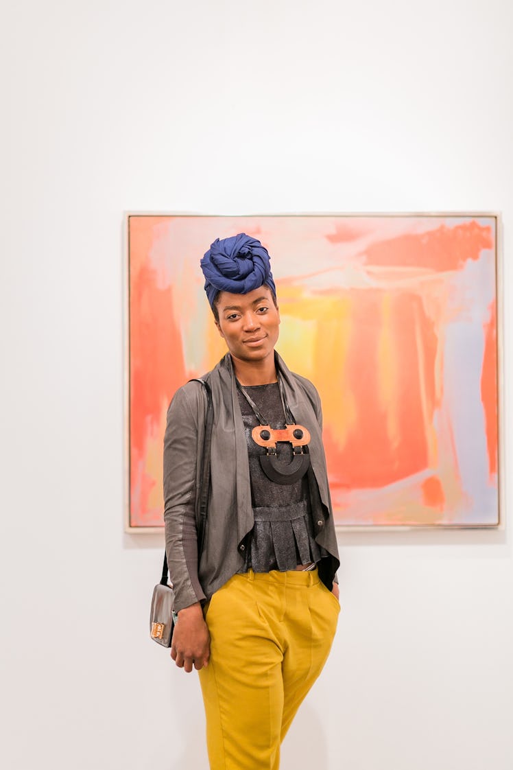 A woman wearing a blue turban, yellow pants, grey top and a jacket standing in front of a painting a...