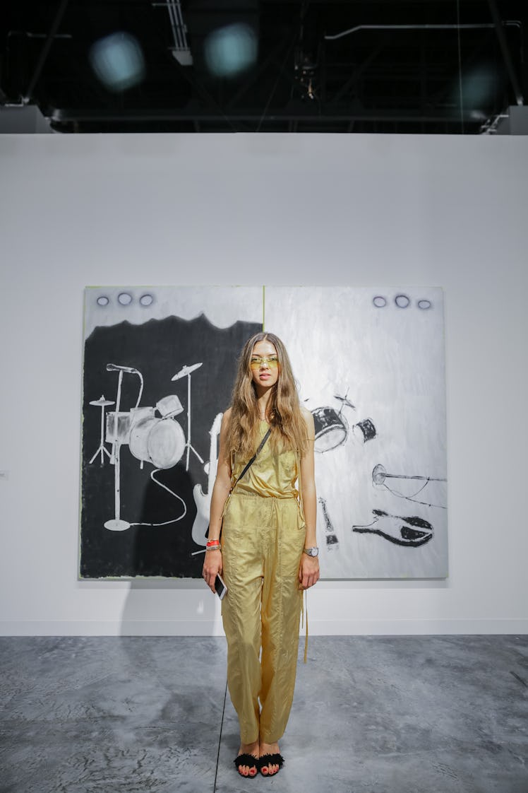 A woman wearing a jumpsuit in golden hue standing in front of a painting at Art Basel.