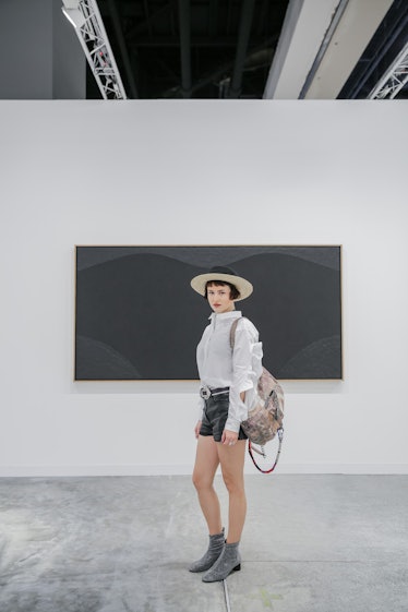 A woman wearing a hat, a white shirt, shorts, and boots posing in front of a painting in black hues ...