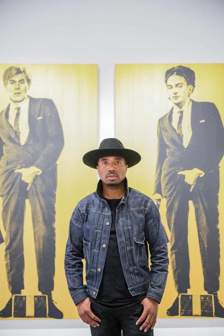 A man in a stylish ensemble of a black hat, t-shirt, pants, and a denim jacket, posing in front of t...