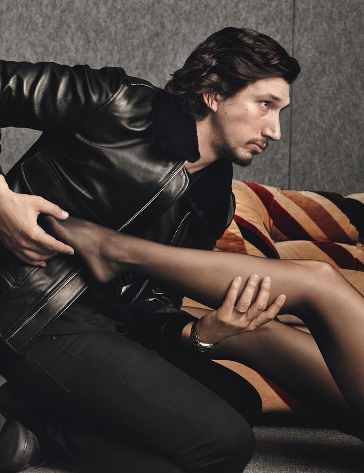 Adam Driver in a black leather jacket and pants leaning forward while holding onto a female leg