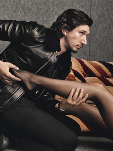 Adam Driver in a leather jacket leaning forward while holding the leg of a woman wearing black tight...