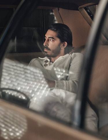 Dev Patel in a beige shirt sitting in the back of a car with brown seats
