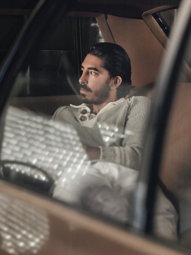 Dev Patel in a beige sweater sitting in the backseat of a car and posing