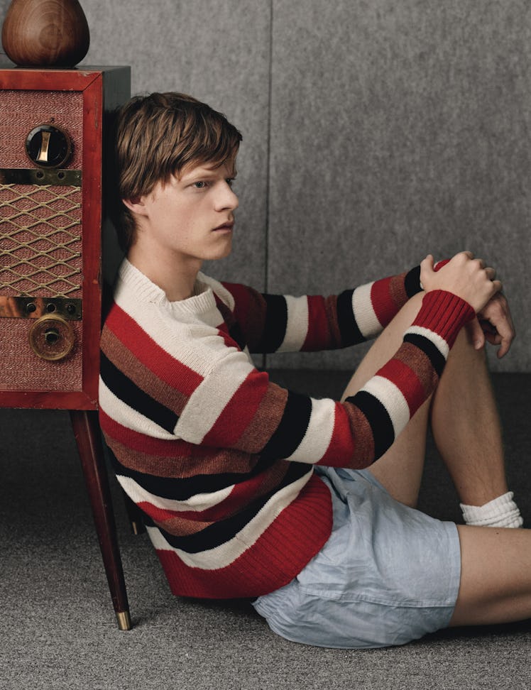 Lucas Hedges sitting next to a table in a red-white striped sweater and blue shorts