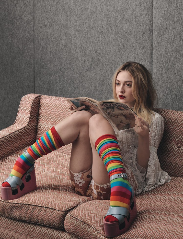 Dakota Fanning in a blue shirt and brown floral short and rainbow striped socks sitting on a couch a...