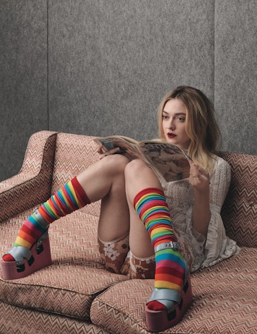 Dakota Fanning in a blue shirt and brown floral short and rainbow striped socks sitting on a couch a...