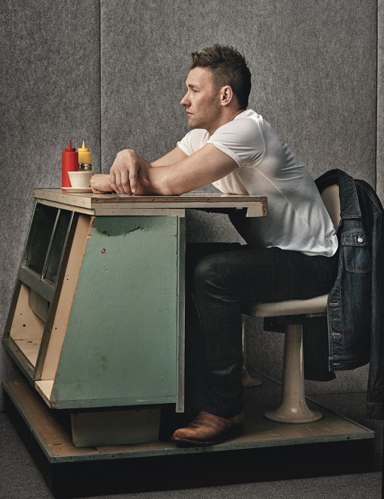 Joel Edgerton in a white shirt and black trousers sitting next to a table
