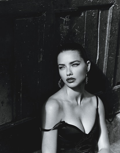 Adriana Lima sitting in a black dress looking into the distance over her right shoulder
