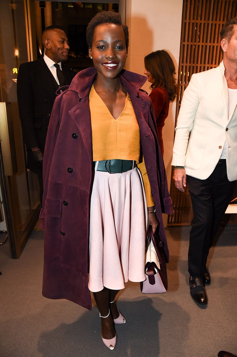 Lupita Nyong’o wearing a white skirt, yellow shirt, and a burgundy coat