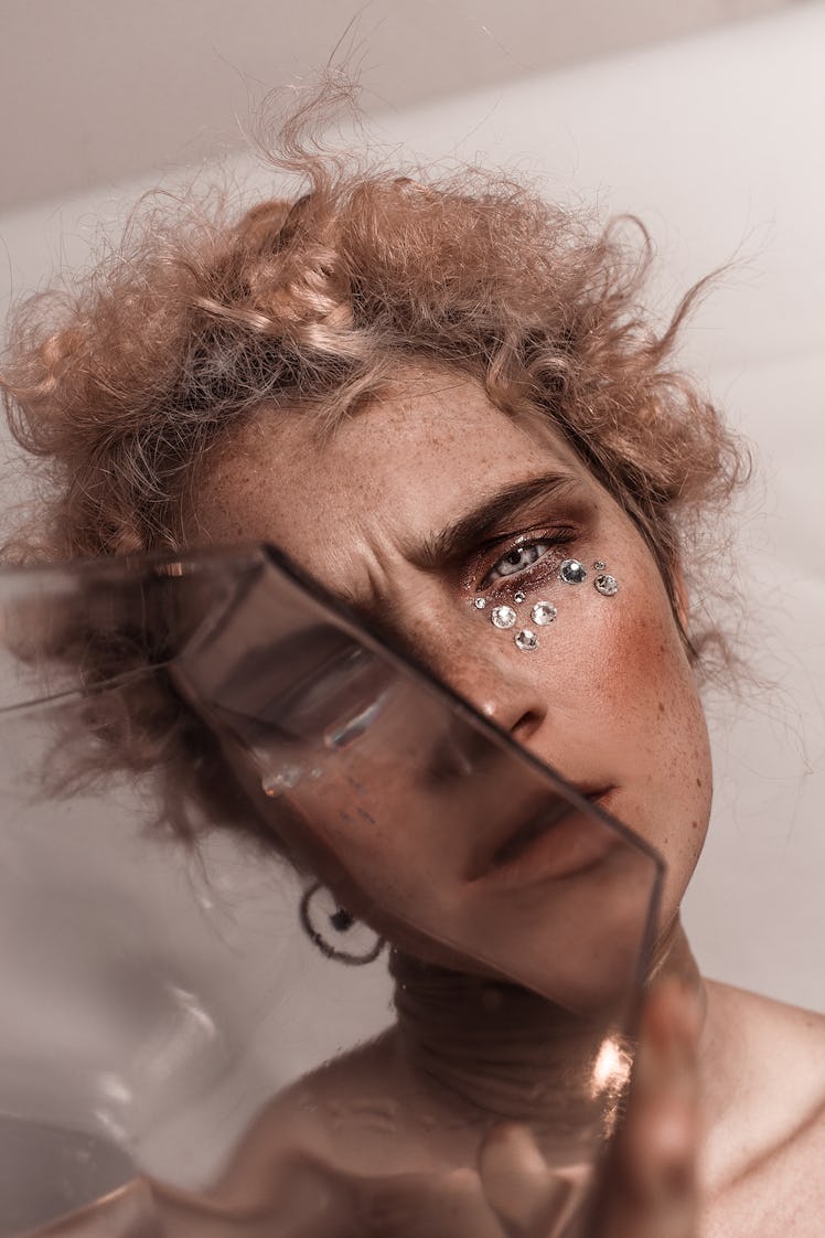 Red-haired model peeking behind a piece of glass, with rhinestones and freckles on her face for In t...