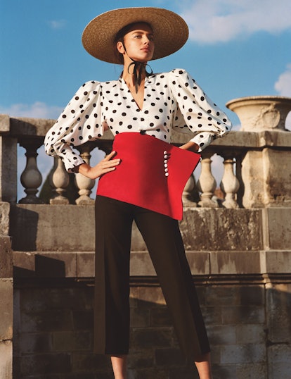 A model wearing a shirt with dots, black pants, a red corset belt, and a straw hat