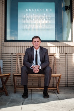 David Colbert in a black suit, white shirt and a black tie sitting on a wooden bench in front of a C...