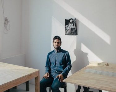 A man sitting in a blue denim shirt and jeans n a room full of light and two desks by Daniel Dorsa