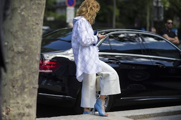 A woman in a blue shirt and white trousers typing on her phone next to a vehicle by Asia Typek