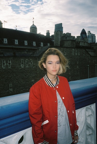 A blonde woman in a red varsity jacket and a grey shirt leaning against a wall by Gunner Stahl