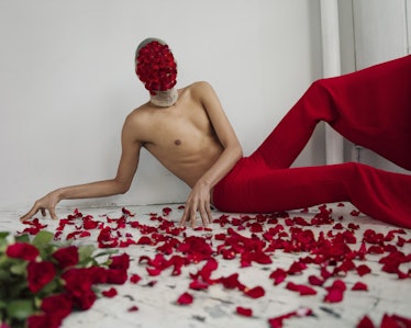 A topless man posing in red trousers with rose petals covering his face and the floor by Matthew Pri...