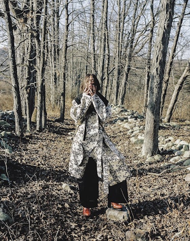 A woman posing in a camo print coat and black trousers in a forest by Julien Boudet