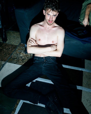 A man sitting on the floor with jeans and no top on, crossing his arms 