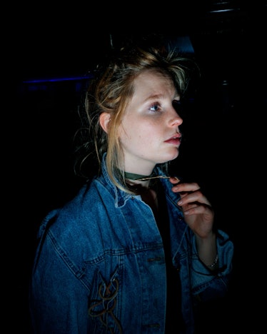 A blonde young woman in a denim jacket looking up while holding on to her necklace 