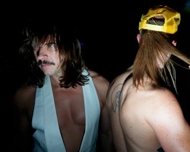 A man in a white vest, with a moustache and shoulder-length hair looking to the side at Ova the Rain...