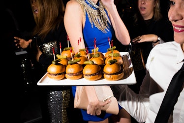 A waiter in a white shirt and black tie handing out mini sliders on a square plate 