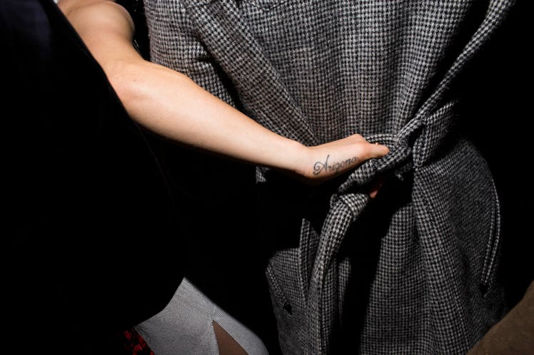 The back of a hand with a tattoo on the side holding onto the belt of a grey coat worn by another pe...
