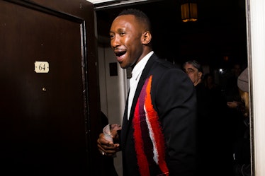 Mahershala Ali in a black blazer with red, orange and purple stripes, and a white shirt laughing 