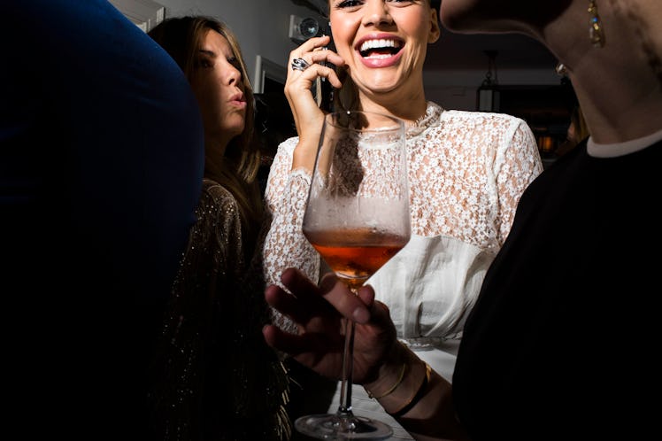 A woman in a white blouse with sequins on top, talking on the phone and laughing while holding a dri...
