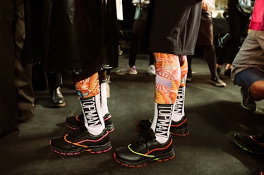 Two male models standing while wearing black shoes, white socks, orange leggings, and black shorts