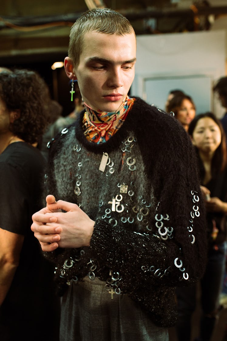 A male model wearing a black shirt and a green cross-shaped earring on his right ear