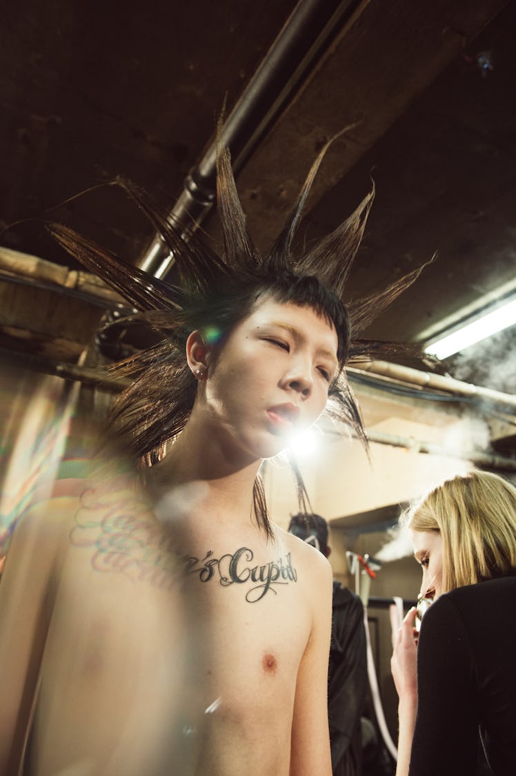 A male model with a rock hairstyle and chest tattoo getting prepared for Charles Jeffrey’s MAN Fall ...