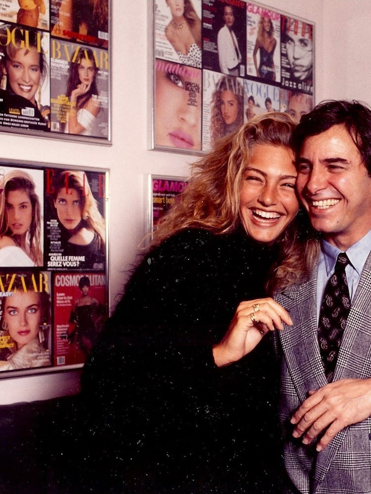John Casablancas standing with a model in front of a wall of magazine covers