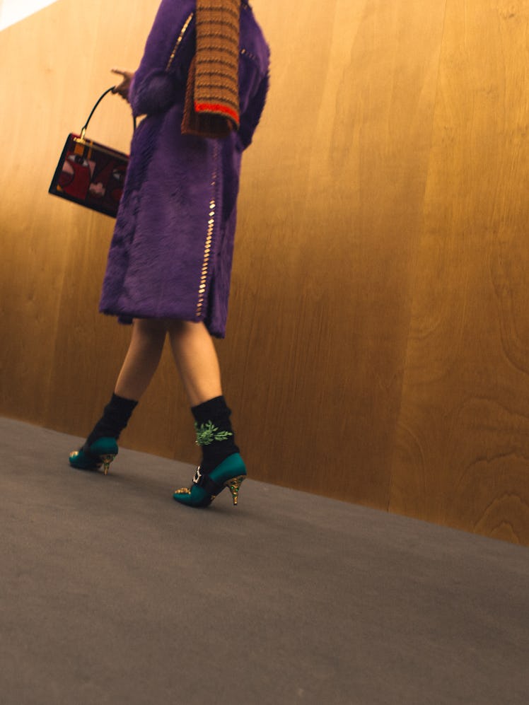 A model in a purple coat walking next to a brown wall backstage at the Fall 2017 Prada Fashion Show