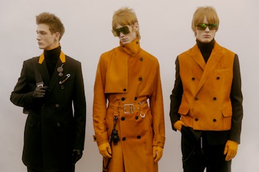 A model in a black coat with an orange collar standing next to models in an orange trench coat and v...