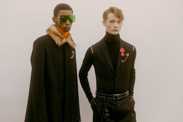 A model in a black coat with a fur collar standing next to a model in a black turtleneck and vest 