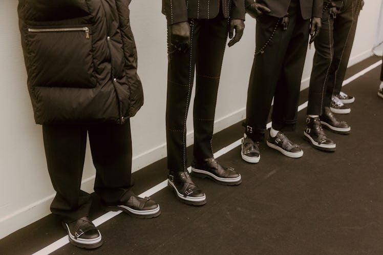 Four models lining up backstage all in black and white shoes, black pants and one has a black puffer...