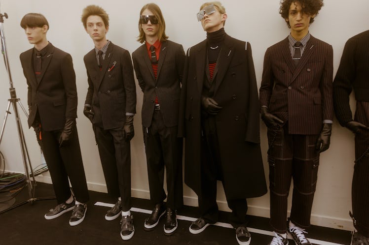 Models lining up backstage at the Dior show: all are in black suits while one is in a turtleneck and...