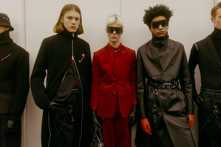 A model in a red suit and two models in black coats standing backstage at the Dior Homme Fall 2017 s...